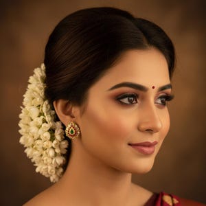 May include: A close-up of a person wearing a gold-colored stud earring with a red and green gemstone center. The earring is surrounded by small white stones. The person has dark hair and is wearing makeup. A flower arrangement is in the hair.