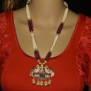 Puede incluir: Un collar de oro con un gran colgante que presenta dos pavos reales con piedras rojas y verdes. El collar está hecho de cuentas blancas y burdeos.