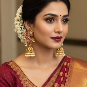May include: Close-up of a woman wearing ornate gold earrings with red and white accents. The earrings feature a peacock design and a bell shape with dangling pearls. The woman is wearing a maroon sari with gold embroidery.