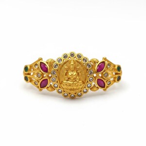 May include: A gold-toned bracelet featuring a central circular medallion with a relief of a seated deity. The medallion is surrounded by small clear stones, flanked by marquise-cut pink stones, and accented with small green stones on the outer edges.
