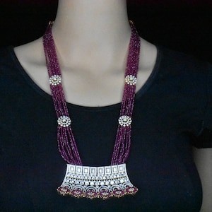 Puede incluir: Un collar largo y de varias capas con un gran colgante adornado. El collar está hecho de pequeñas cuentas moradas y presenta un colgante plateado con piedras transparentes.