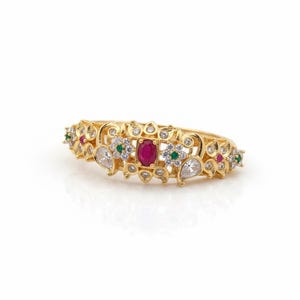 May include: A gold ring featuring a central oval-cut red gemstone flanked by small green and clear stones. The band is decorated with intricate metalwork, including floral motifs and additional clear stones arranged in a symmetrical pattern.