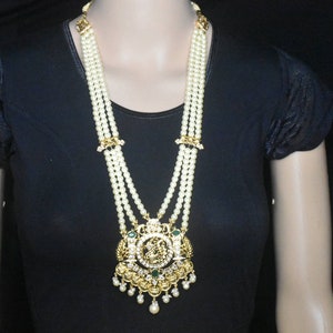 Puede incluir: Un collar de perlas de varias hileras con un colgante de oro que presenta una piedra verde y detalles intrincados. El collar está en un maniquí que lleva una camisa negra.