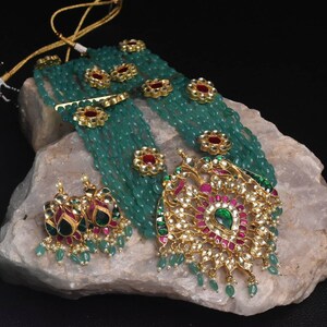 May include: A multi-strand necklace with green beads and gold accents. The necklace features a large pendant with a floral design and red and green gemstones. The pendant is attached to a chain with a green bead tassel. The necklace is displayed on a rock.