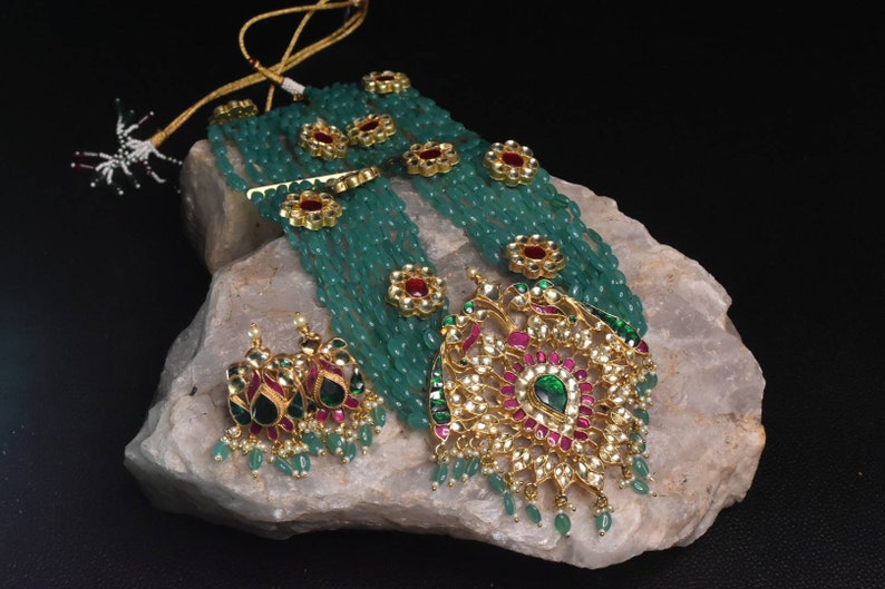 May include: A multi-strand necklace with green beads and gold accents. The necklace features a large pendant with a floral design and red and green gemstones. The pendant is attached to a chain with a green bead tassel. The necklace is displayed on a rock.
