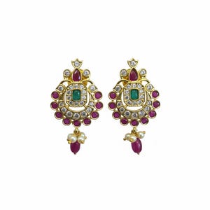 May include: A pair of gold-coloured earrings with a floral design. Each earring features a central square green stone, surrounded by white and red stones. Small white pearls and a red stone dangle from the bottom.