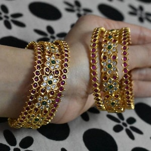 Può includere: Un set di cinque braccialetti d'oro con intricati disegni floreali. I braccialetti sono adornati con pietre preziose rosa, bianche e verdi.