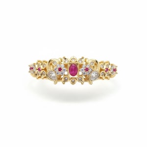 May include: A gold ring featuring a central oval-cut red gemstone surrounded by small clear stones. The band is decorated with floral motifs, additional red stones, and pear-shaped clear stones arranged in a symmetrical, ornate metal pattern.