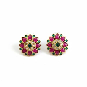 May include: A pair of gold-toned stud earrings. Each earring features a floral design with a central green stone surrounded by small white stones. The petals are a deep pink color, and the outer edge has green accents.