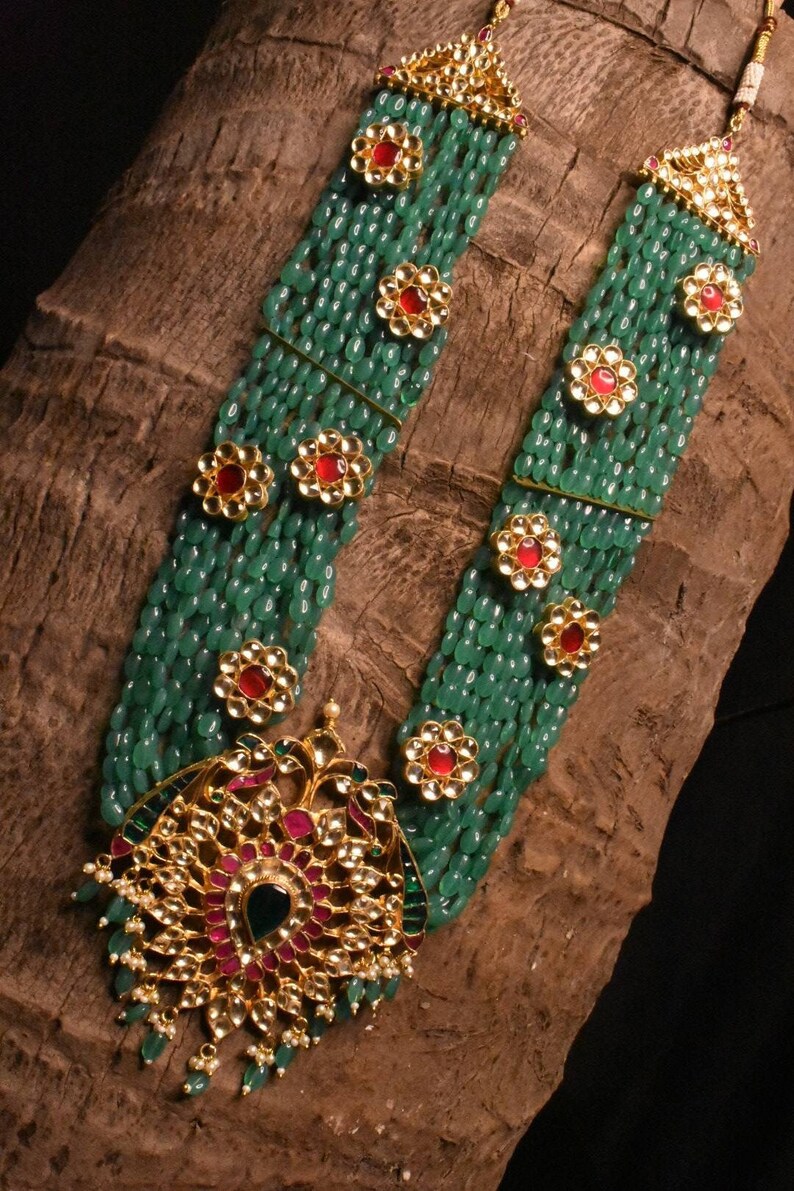 May include: A long, multi-strand necklace with green beads and gold accents. The necklace features a large, ornate pendant with red and green stones.