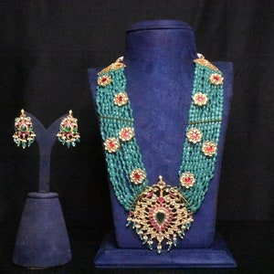 May include: A multi-strand necklace with a large, ornate pendant featuring a peacock design. The necklace is made of turquoise beads and gold accents. The pendant is decorated with red and green gemstones. The necklace is displayed on a blue velvet stand. Matching earrings with green and red gemstones are also included.