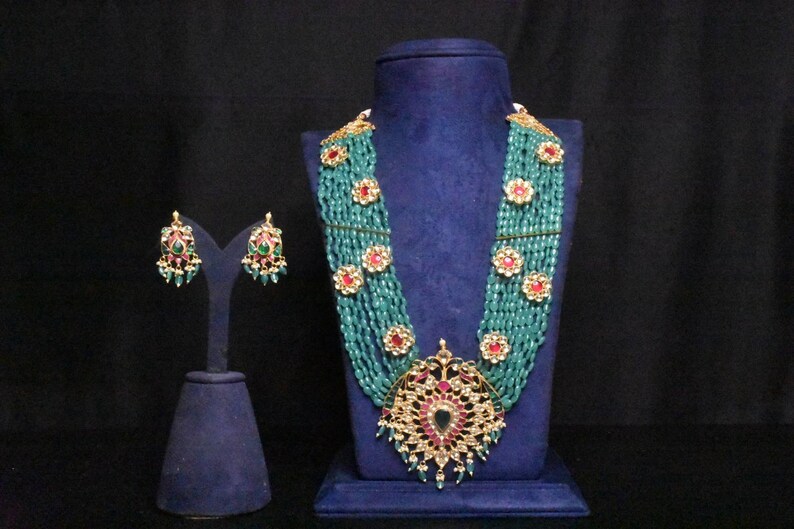 May include: A multi-strand necklace with a large, ornate pendant featuring a peacock design. The necklace is made of turquoise beads and gold accents. The pendant is decorated with red and green gemstones. The necklace is displayed on a blue velvet stand. Matching earrings with green and red gemstones are also included.