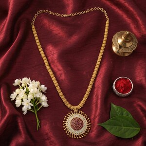 May include: A gold-toned necklace featuring a long chain with textured spherical links and a large, circular pendant. The pendant has a central cluster of small red stones surrounded by a ring of clear stones and radiating gold rays.