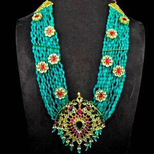 May include: A multi-strand necklace made of turquoise beads with gold accents and red and green gemstones. The necklace has a large, ornate pendant with a floral design.