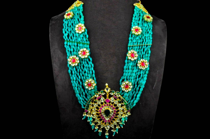 May include: A multi-strand necklace made of turquoise beads with gold accents and red and green gemstones. The necklace has a large, ornate pendant with a floral design.