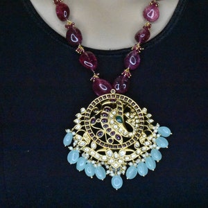 May include: A gold necklace with a large pendant featuring a peacock design. The pendant is adorned with red, green, and white gemstones. The necklace is strung with red gemstone beads.