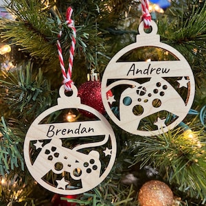 May include: Two wooden Christmas ornaments shaped like a circle with a cutout of a video game controller. The ornaments are decorated with stars and have the names "Andrew" and "Brendan" written on them.