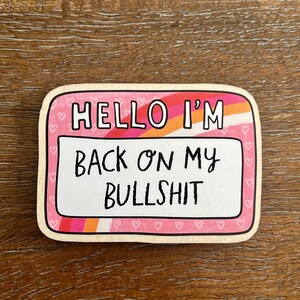 May include: Pink and white name tag sticker with the text "Hello I'm Back On My Bullshit" in black lettering. The sticker has a rainbow stripe and a border of pink hearts.