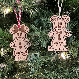 May include: Two wooden gingerbread person ornaments with a bow and a bow tie. The ornaments are hanging on a Christmas tree. The ornament on the left says "Luna" and the ornament on the right says "Brendan".