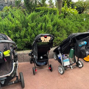 May include: Four black strollers are lined up in a row. The strollers have black fabric seats and black frames. The strollers have various accessories attached to them, including a green water bottle, a black bag, and a Mickey Mouse themed wooden sign.