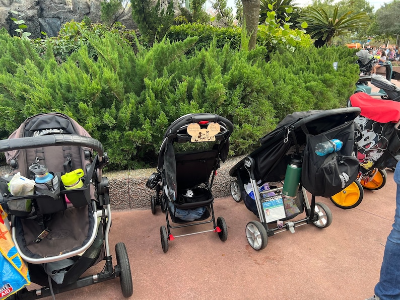 May include: Four black strollers are lined up in a row. The strollers have black fabric seats and black frames. The strollers have various accessories attached to them, including a green water bottle, a black bag, and a Mickey Mouse themed wooden sign.