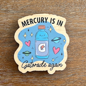 Puede incluir: Una pegatina de madera con fondo azul y una ilustración de dibujos animados de una botella de Gatorade azul con una cara triste. El texto "MERCURY IS IN Gatorade again" está escrito en negro en la pegatina.