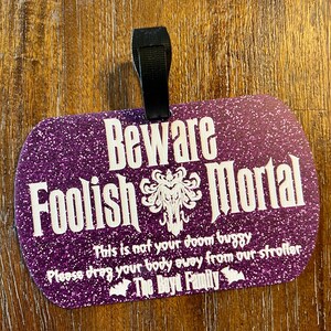 May include: Purple glitter luggage tag with the text "Beware Foolish Mortal" and a spooky graphic. The tag also includes the text "This is not your doom buggy. Please drag your body away from our stroller. The Boyd Family."
