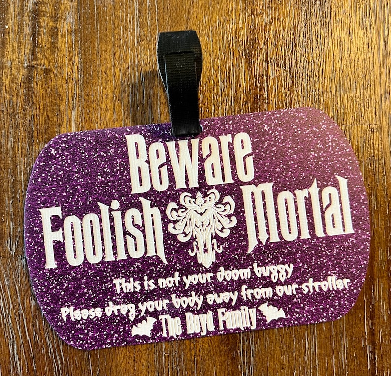May include: Purple glitter luggage tag with the text "Beware Foolish Mortal" and a spooky graphic. The tag also includes the text "This is not your doom buggy. Please drag your body away from our stroller. The Boyd Family."