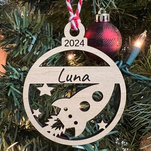 Peut inclure: Une décoration de Noël en bois en forme de cercle avec une fusée et des étoiles à l'intérieur. La décoration est personnalisée avec le nom "Luna" et l'année "2024".