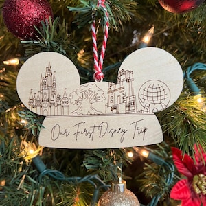 May include: A wooden ornament shaped like Mickey Mouse ears with a laser-engraved image of Disney World landmarks and the text "Our First Disney Trip".