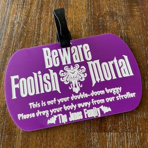 May include: A purple Halloween-themed sign with the text "Beware Foolish Mortal" and a spooky graphic. The sign also includes the text "This is not your double-doom buggy. Please drag your body away from our stroller. The Jones Family."