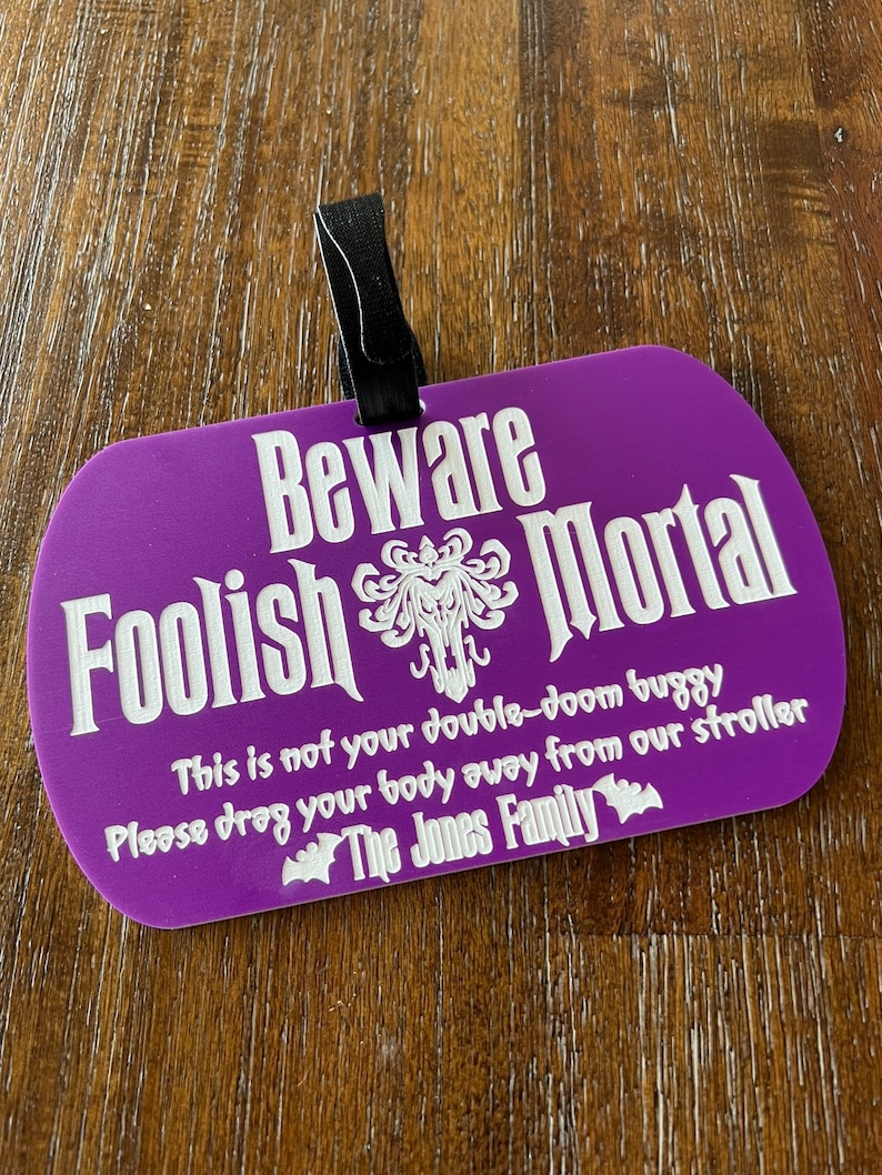 May include: A purple Halloween-themed sign with the text "Beware Foolish Mortal" and a spooky graphic. The sign also includes the text "This is not your double-doom buggy. Please drag your body away from our stroller. The Jones Family."