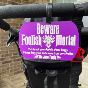 May include: A purple sign with white text that reads "Beware Foolish Mortal" with a spooky graphic. The sign is attached to a black stroller with the text "This is not your double-doom buggy. Please drag your body away from our stroller. The Jones Family."
