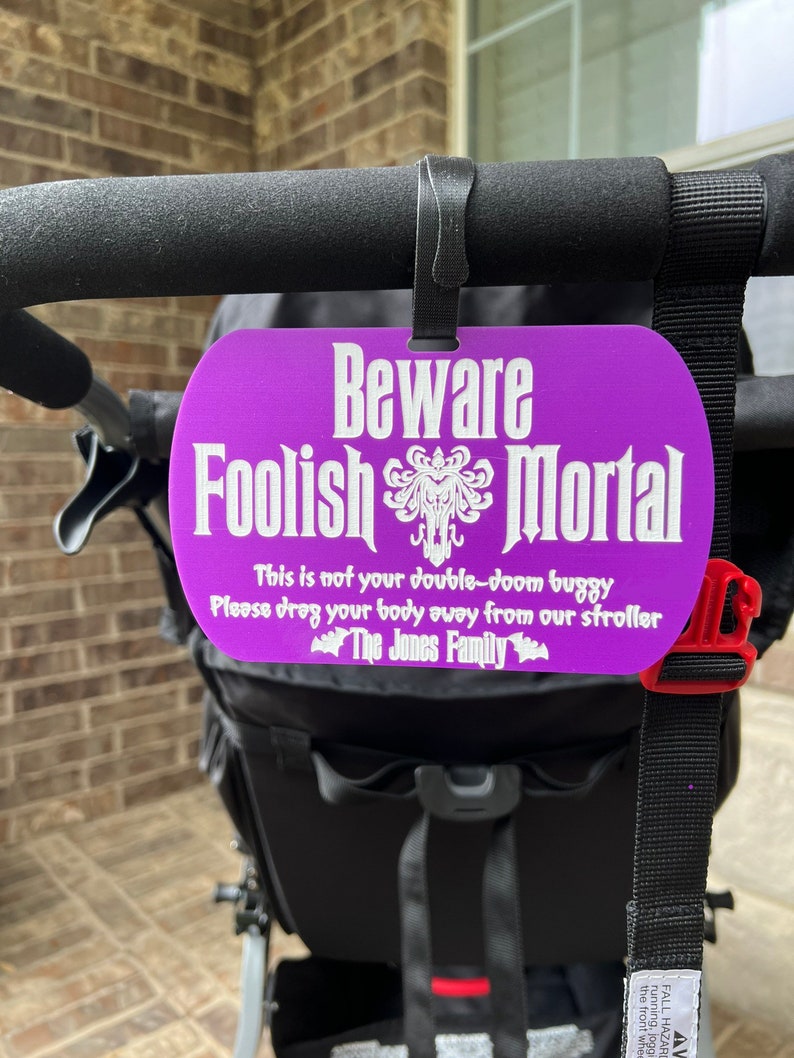 May include: A purple sign with white text that reads "Beware Foolish Mortal" with a spooky graphic. The sign is attached to a black stroller with the text "This is not your double-doom buggy. Please drag your body away from our stroller. The Jones Family."