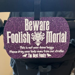 May include: Purple glitter stroller tag with the text "Beware Foolish Mortal" and a white silhouette of a ghost. The text below reads "This is not your doom buggy. Please drag your body away from our stroller. The Boyd Family."