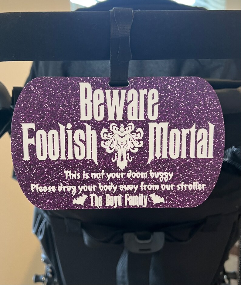 May include: Purple glitter stroller tag with the text "Beware Foolish Mortal" and a white silhouette of a ghost. The text below reads "This is not your doom buggy. Please drag your body away from our stroller. The Boyd Family."