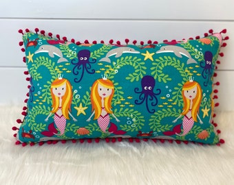 May include: Rectangular teal pillow with a mermaid and sea creature pattern. The design includes mermaids, dolphins, octopuses, and starfish. The pillow is trimmed with a vibrant red pom-pom fringe. A decorative accent for a nautical or fantasy-themed room.