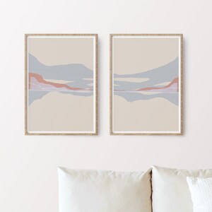 Puede incluir: Dos impresiones abstractas de paisajes enmarcadas con un fondo beige y formas abstractas azules y rosas. Las impresiones est&aacute;n colgadas en una pared blanca sobre un sof&aacute; blanco con un reposapi&eacute;s tejido.
