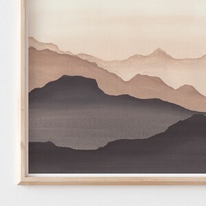 May include: A framed watercolor painting of a mountain range. The artwork features layers of mountains in shades of brown and gray, with a light beige sky. The frame is made of light-colored wood.