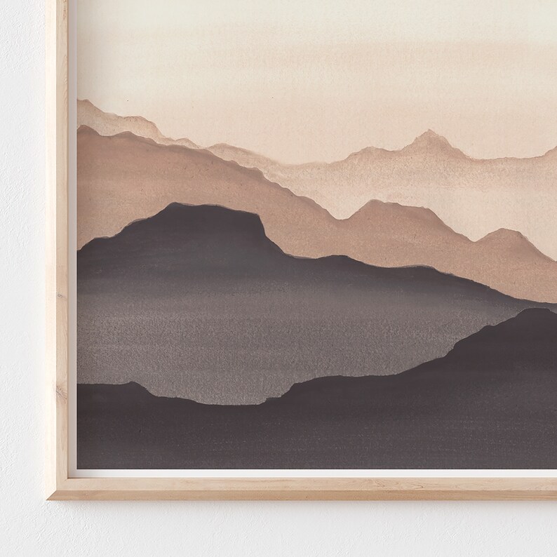 May include: A framed watercolor painting of a mountain range. The artwork features layers of mountains in shades of brown and gray, with a light beige sky. The frame is made of light-colored wood.
