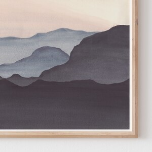 May include: Watercolor landscape art print featuring layered mountain silhouettes in shades of blue and gray, set against a soft pink and beige sky. The artwork is framed in a natural wood frame, adding a touch of warmth to the serene scene.