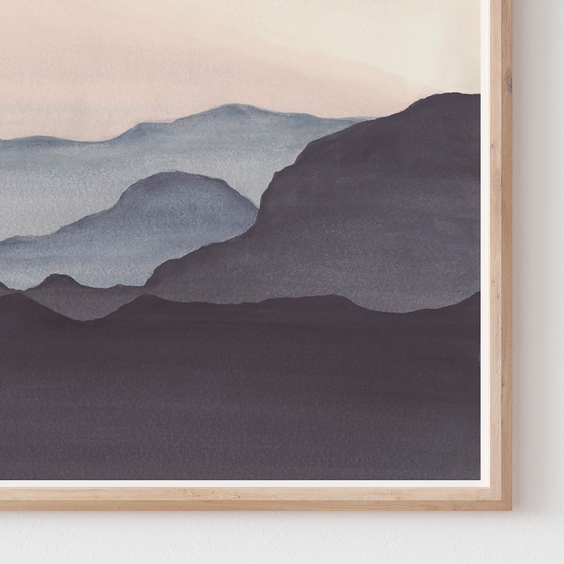 May include: Watercolor landscape art print featuring layered mountain silhouettes in shades of blue and gray, set against a soft pink and beige sky. The artwork is framed in a natural wood frame, adding a touch of warmth to the serene scene.