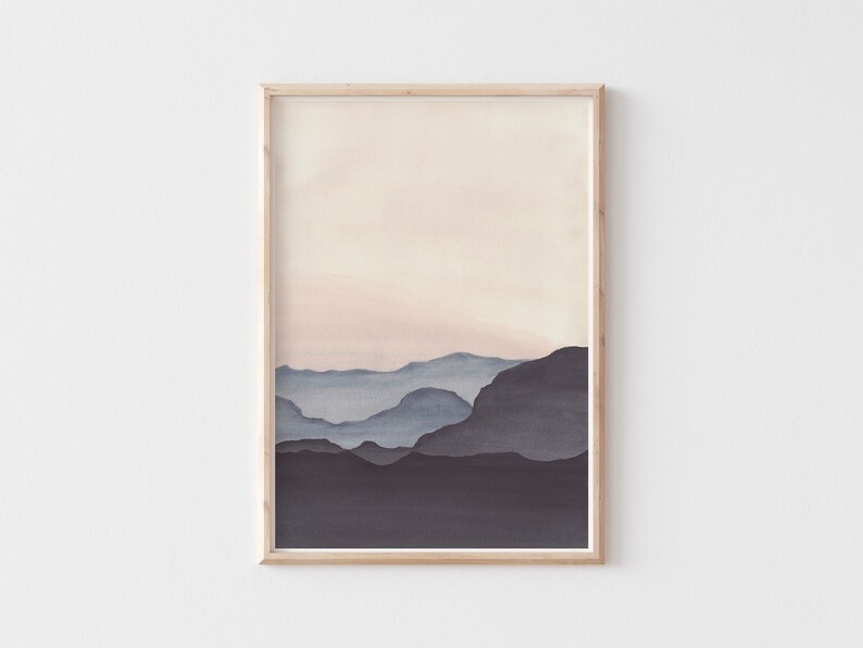 May include: A framed watercolor print featuring a minimalist landscape. The artwork depicts layered mountain silhouettes in shades of blue and gray against a soft, peachy sky. The frame is a light wood color, adding a natural touch to the artwork.