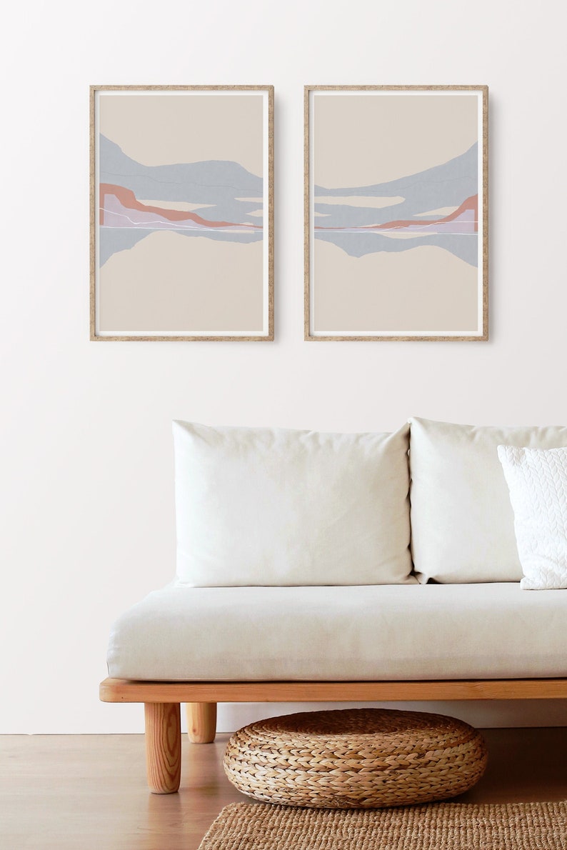 Puede incluir: Dos impresiones abstractas de paisajes enmarcadas con un fondo beige y formas abstractas azules y rosas. Las impresiones est&aacute;n colgadas en una pared blanca sobre un sof&aacute; blanco con un reposapi&eacute;s tejido.