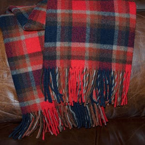 May include: A red, navy, and brown plaid scarf with a fringed edge. The scarf is draped over a brown leather surface. The plaid pattern is a classic design, with a mix of warm and cool colors.