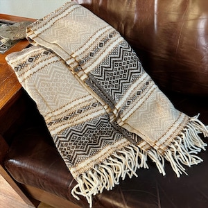 May include: A folded beige and brown woven blanket with geometric patterns and fringe. The blanket features a repeating diamond and zigzag design in shades of brown and white. The blanket is draped over a brown leather surface.