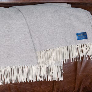 Peut inclure: Une couverture en laine grise avec une frange blanche. La couverture porte une étiquette bleue avec le texte "PENDLETON".