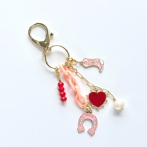 Op de afbeelding: Een gouden sleutelhanger met een roze hoefijzerbedel, een rode hartbedel, een roze laarsbedel, een roze ketting en rode kralen.