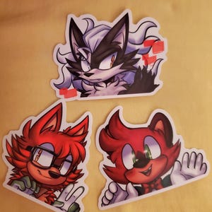 Puede incluir: Tres pegatinas de dibujos animados con animales antropomórficos. Una muestra un lobo blanco y negro con una expresión severa. Las otras dos representan zorros rojos, uno con gafas y el otro saludando. Todas las pegatinas tienen un borde blanco.