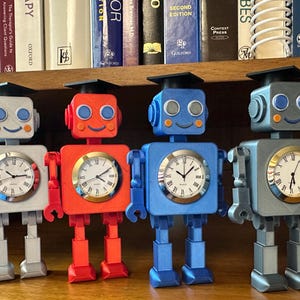 May include: Four colorful robot-shaped clocks with square bodies, round clock faces, and articulated limbs. Each robot wears a graduation cap. Colors include gray, red, blue, and a lighter blue. The clocks are on a wooden surface.